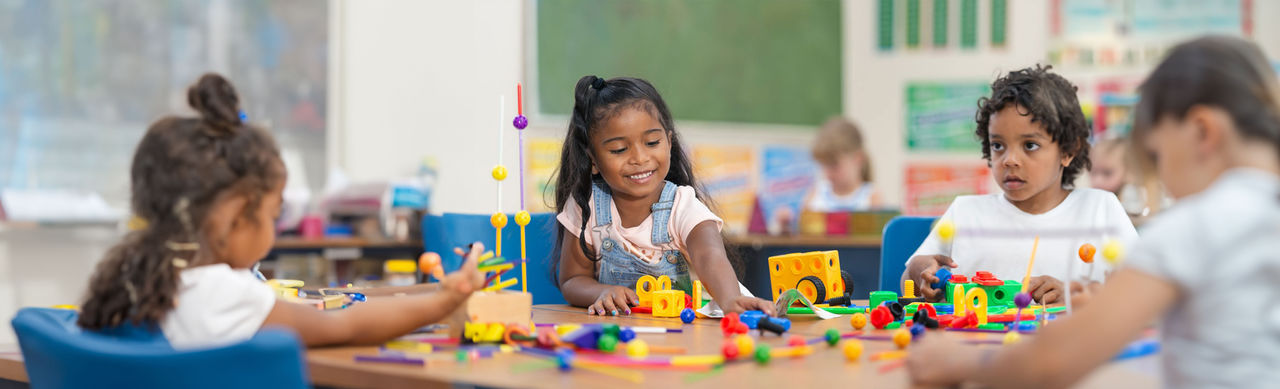 A smiling girl sits at a table in her classroom, building a structure with interlocking pieces.