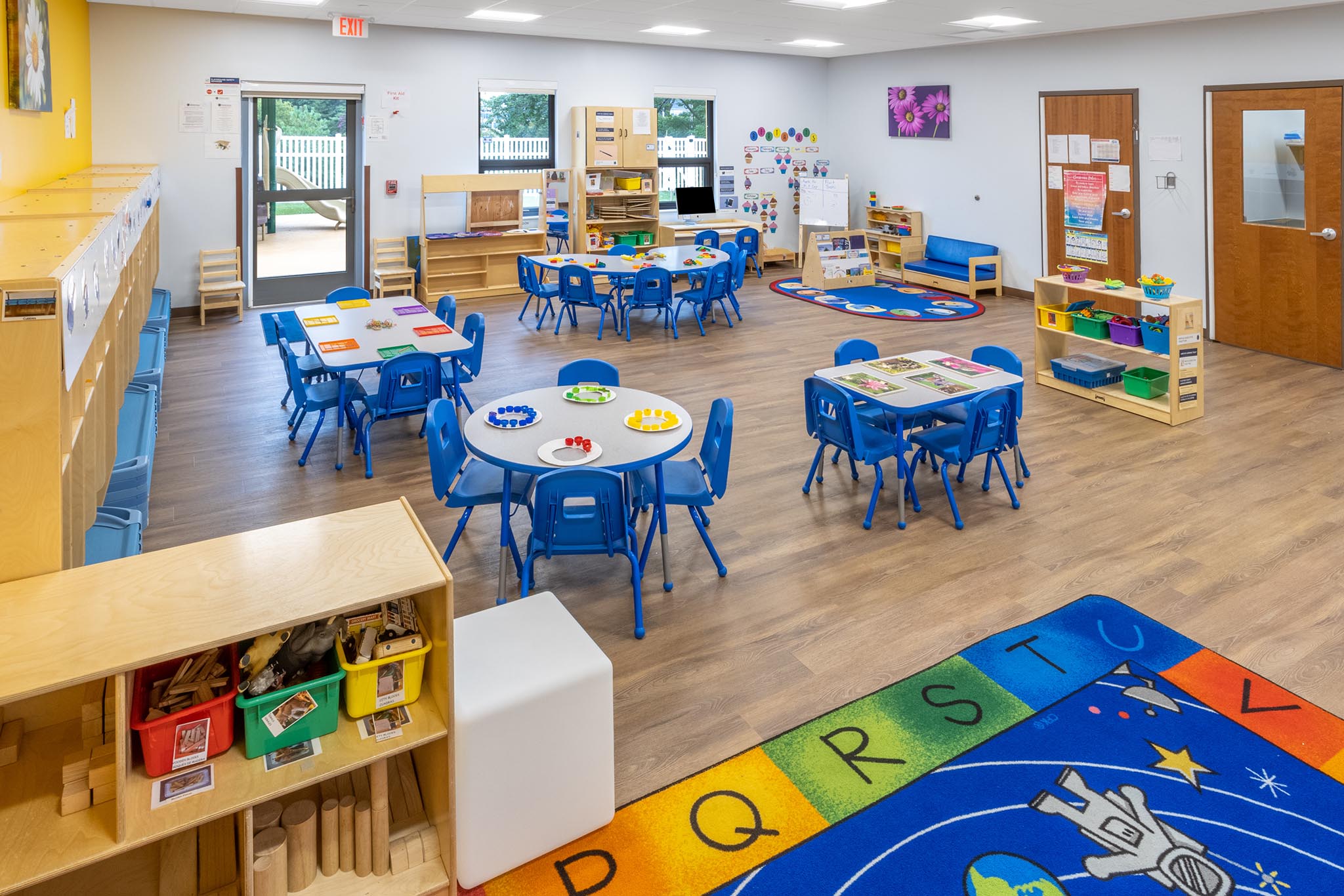 A large Goddard School classroom with a half moon blue table and blue chairs. A green, blue and brown alphabet rug is on the floor near shelves with baskets for storage. 