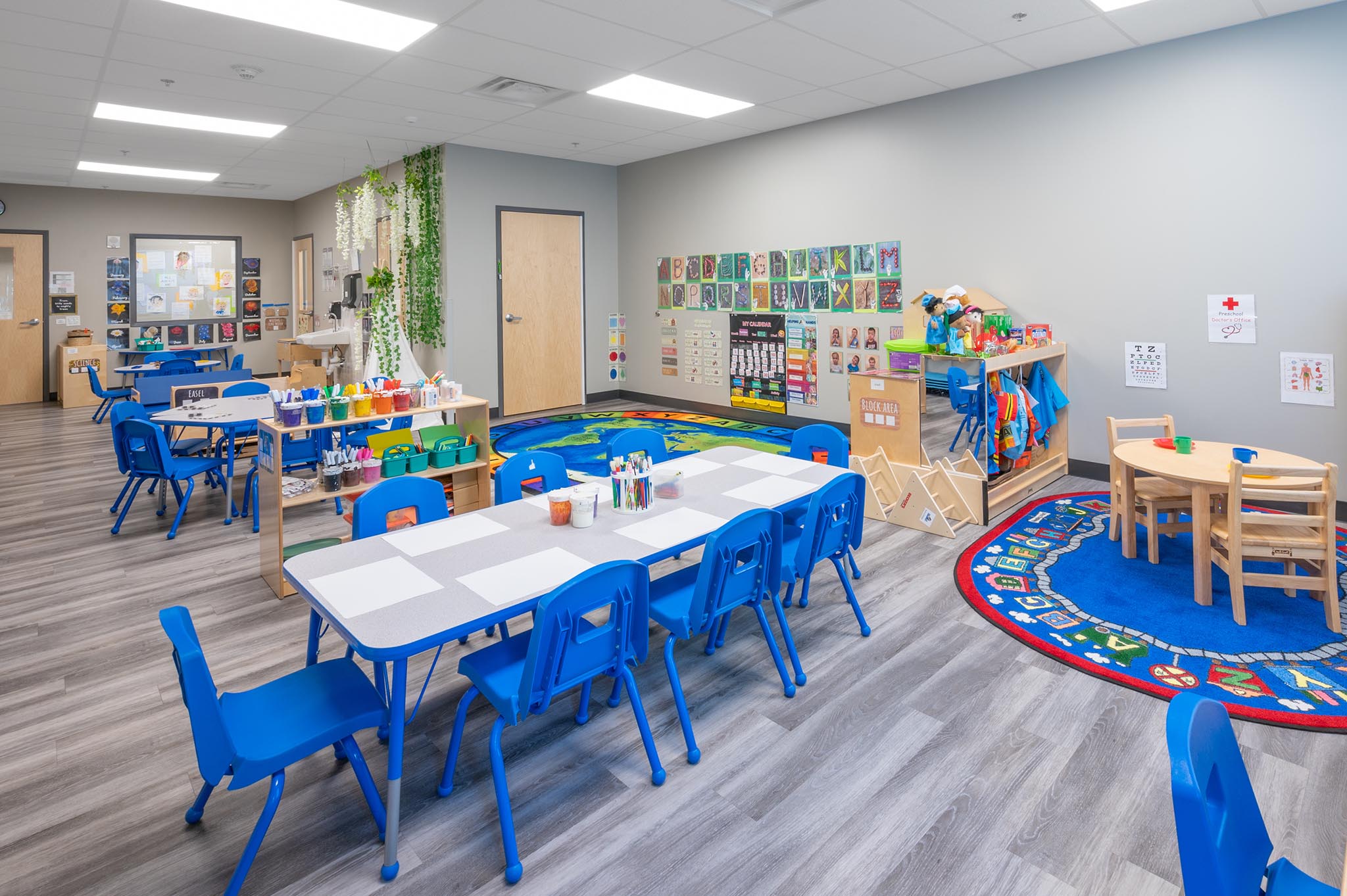 A clean, bright Goddard School classroom with light blue walls, four small tables and green chairs.