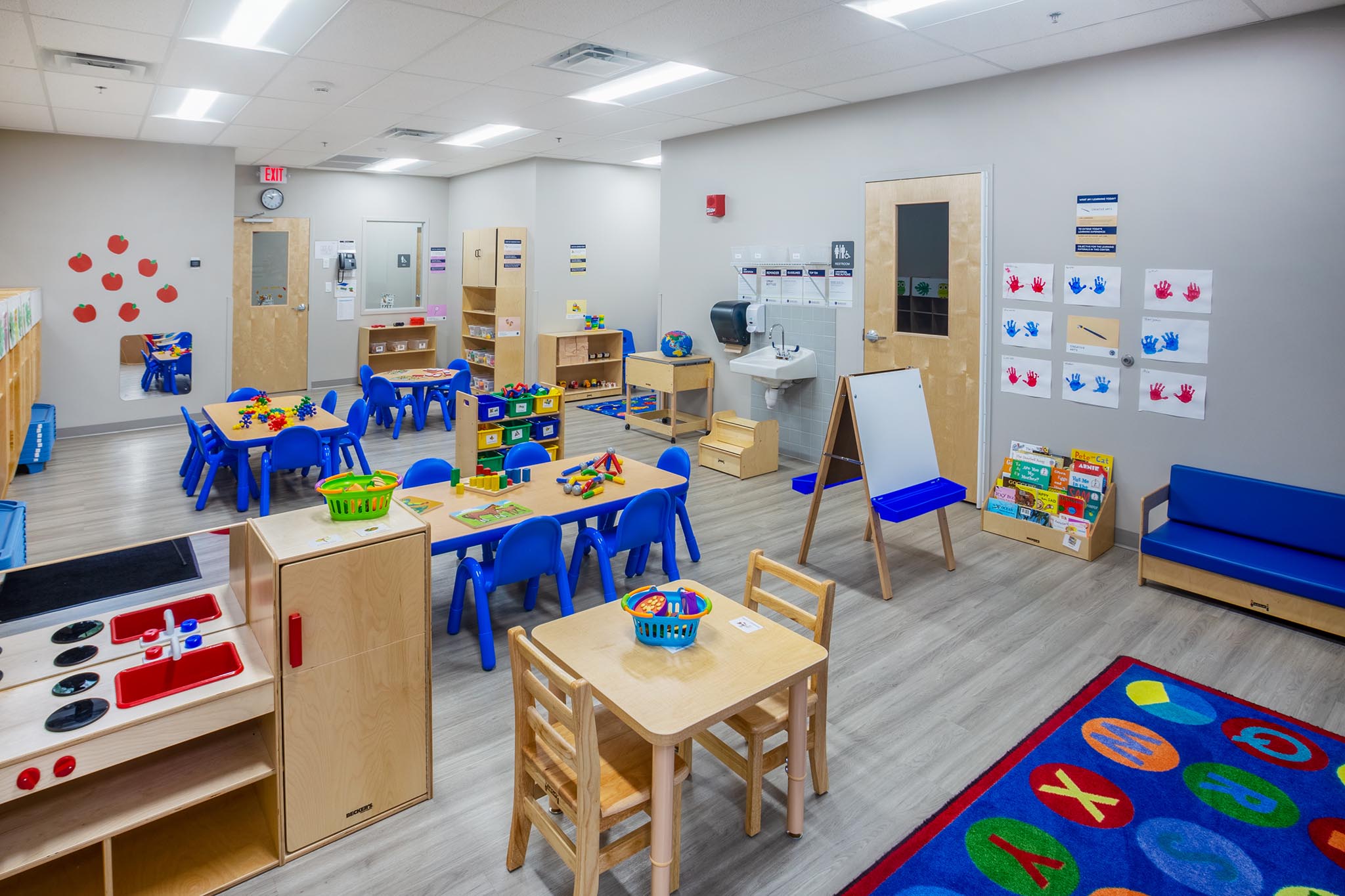 A Goddard School toddler classroom with small blue tables and chairs. Wooden cubbies against the wall, a small wooden play kitchen and a bookshelf with colorful toys.