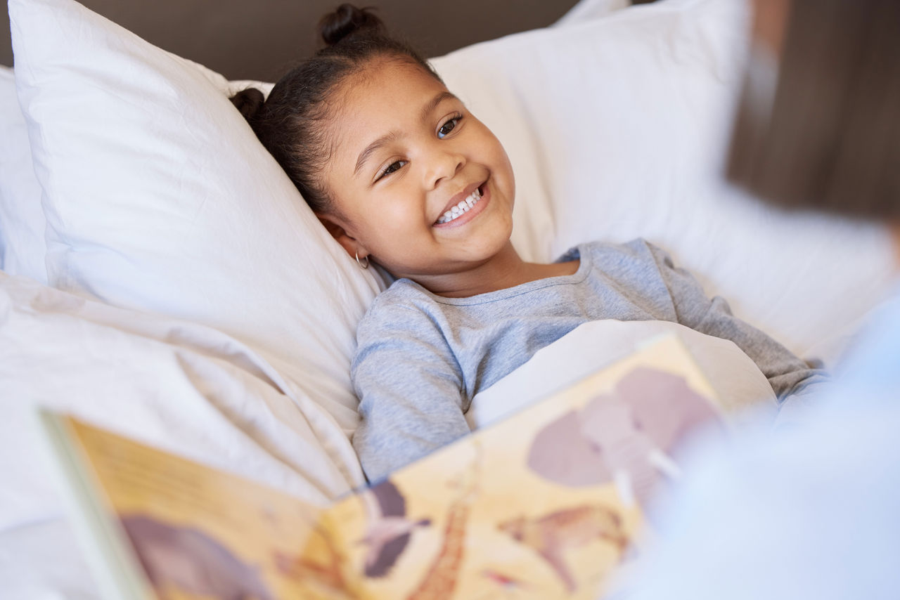 Smiling little girl lying in bed while a parent reads a book