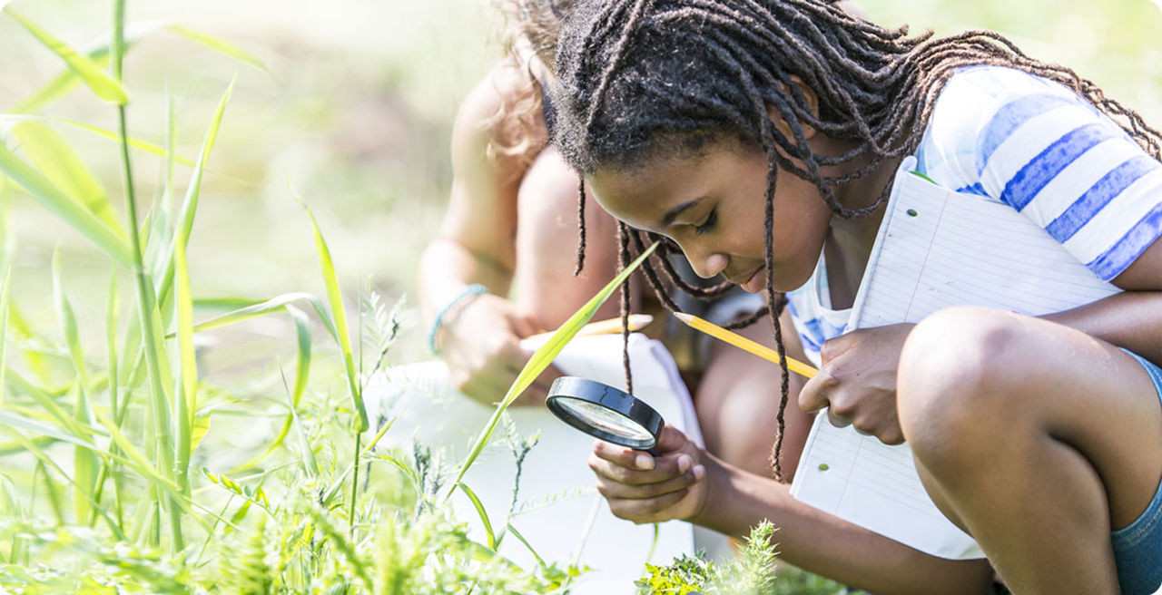 Girl with magnifying glass, notebook and pencil looking at grass