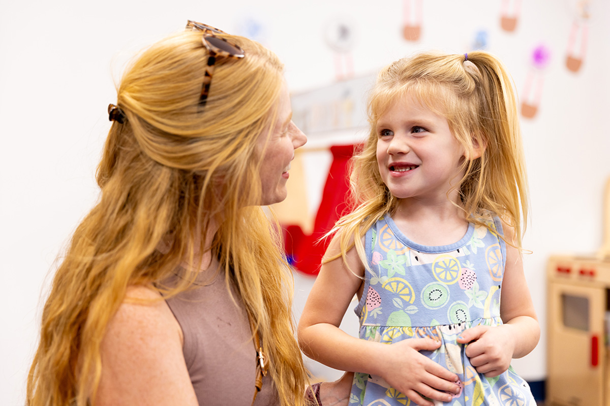 Adult and child interacting in a bright preschool classroom with colorful educational toys and play kitchen in the background.