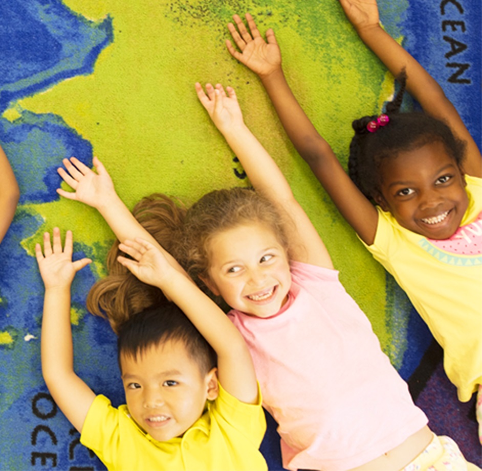 Three diverse children lying on a map rug smiling with hands raised above their heads