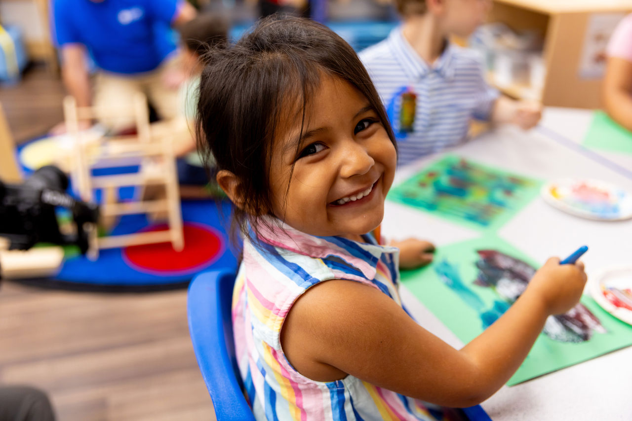A smiling young girl creating an art project at a table in a preschool classroom.