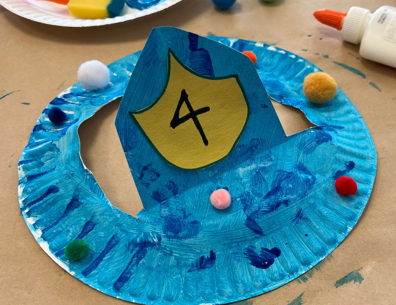 Paper plate fire helmet painted blue with pom poms and the number 4 on the shield
