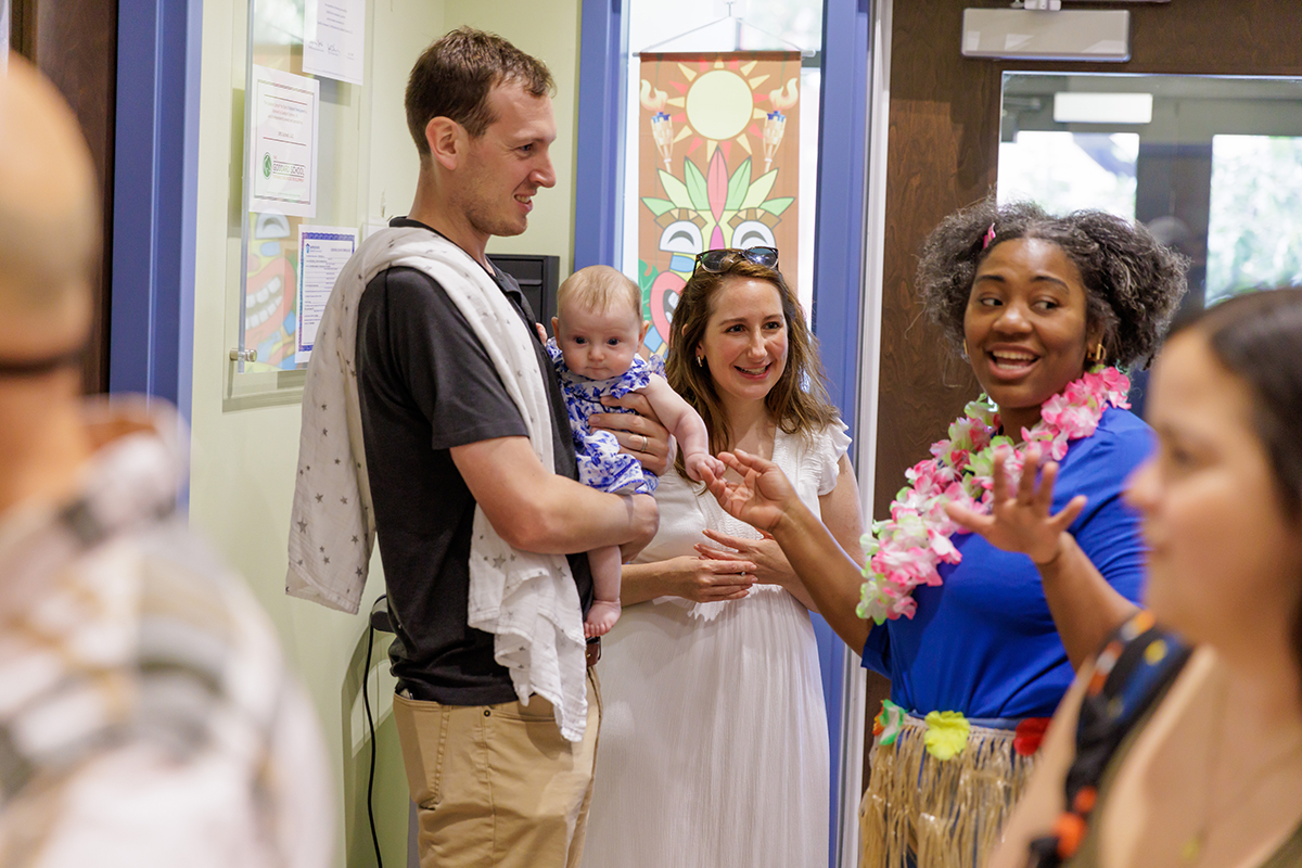 A joyful group of adults and a baby at an indoor gathering. A man holds the baby, while two women, one in a lei, engage in lively conversation.