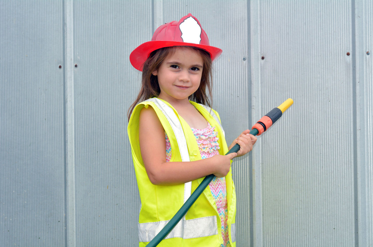 A young girl wearing a safety vest and fire person hat holding a hose