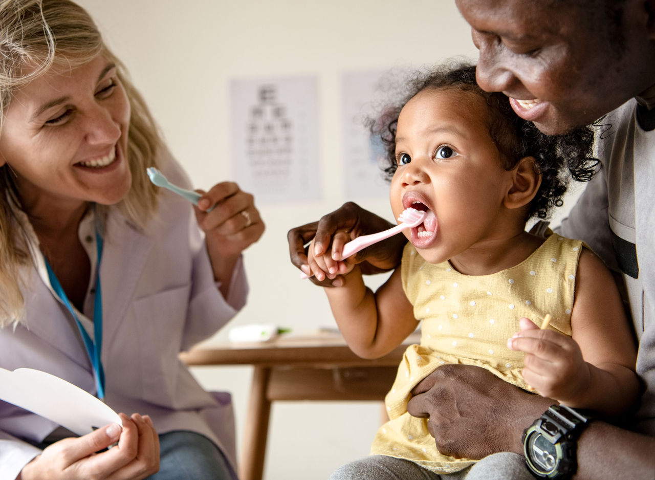 A little girl sitting in her dad's lap holding a toothbrush up to her mouth while a dentist looks on smiling