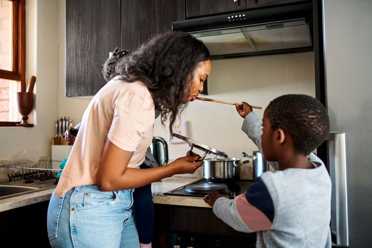 A mother tasting food off a spoon her son is holding up to her at the stove