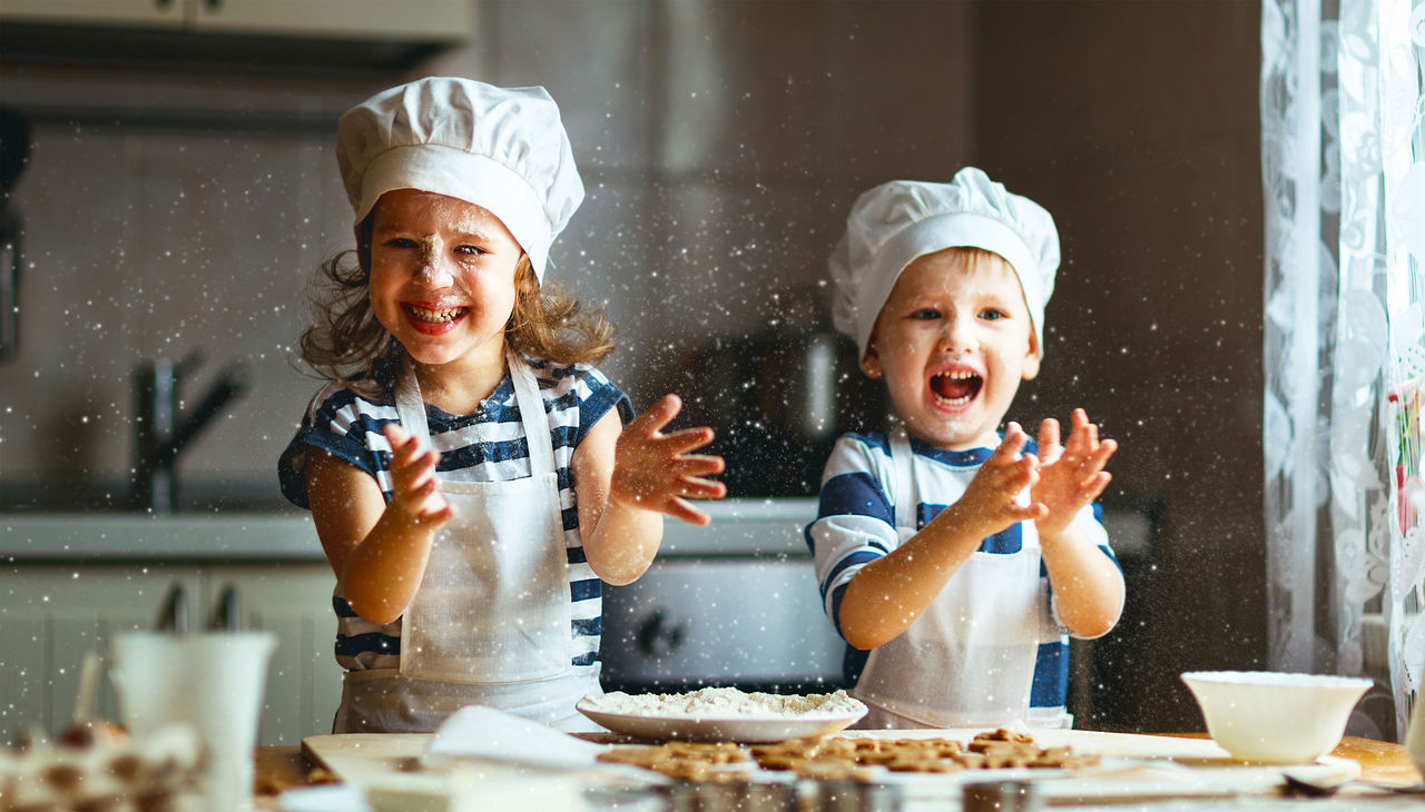 Two children wearing aprons and chef hats clapping their hands in the kitchen
