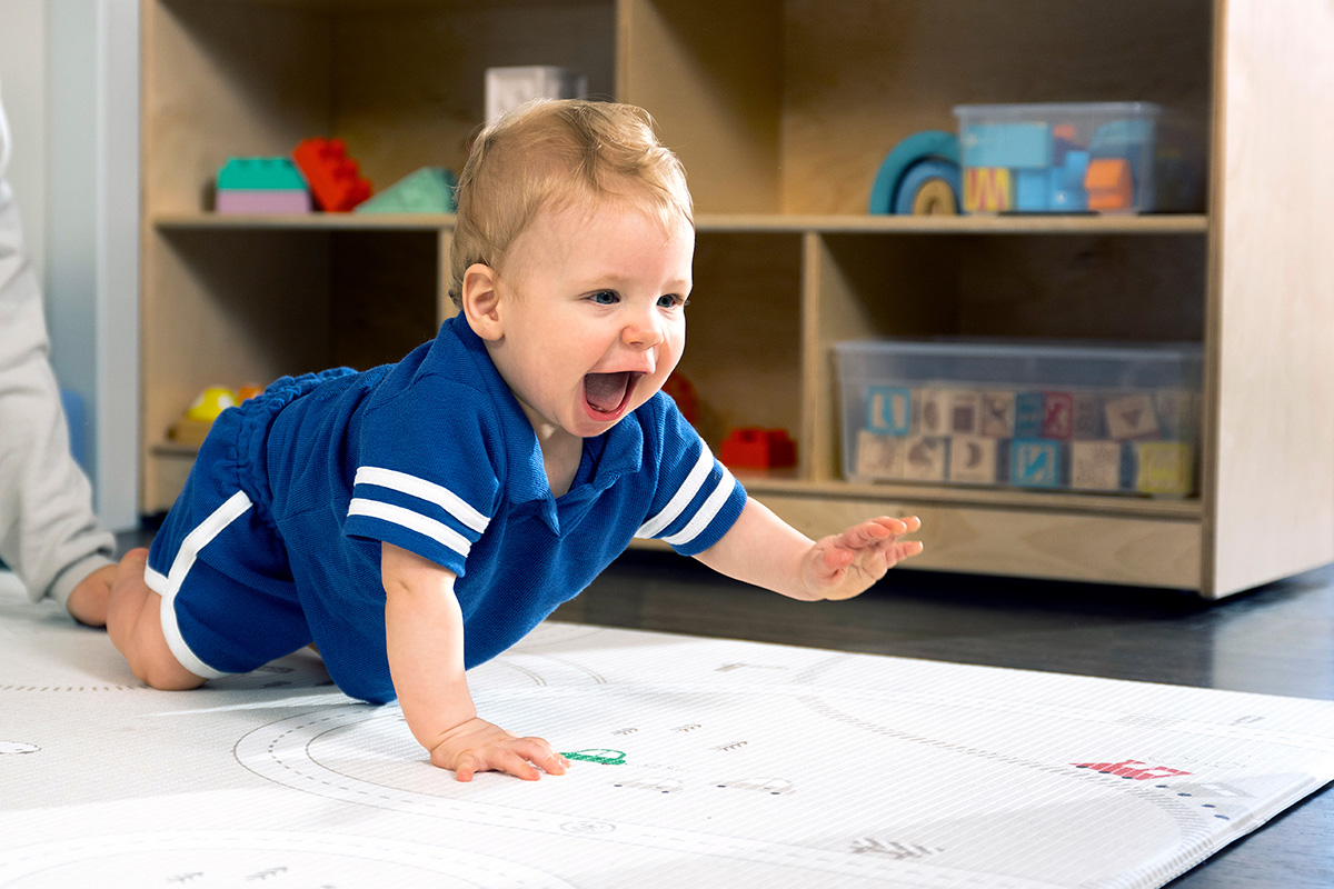 A baby in a blue outfit practicing tummy-time and crawling on a play mat in a brightly lit early learning classroom with shelves of toys in the background.