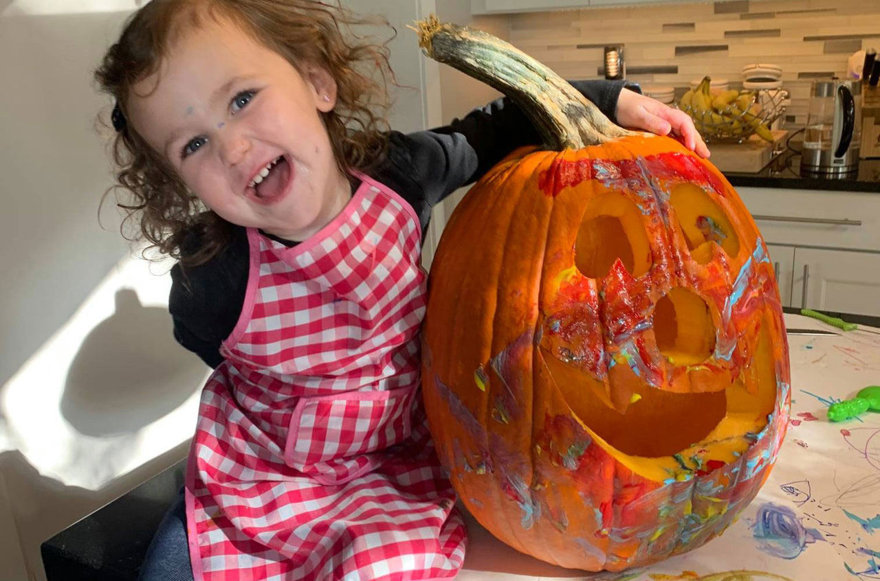 Child wearing a checkered apron smiling next to a pumpkin that she painted