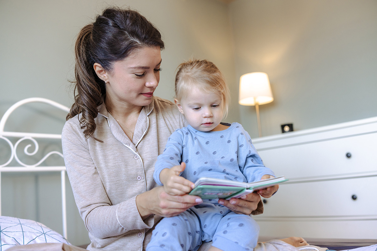 Adult sitting on a bed reading a colorful book to a young child in blue pajamas, with a white metal bed frame and a lit lamp on a nightstand in the background.