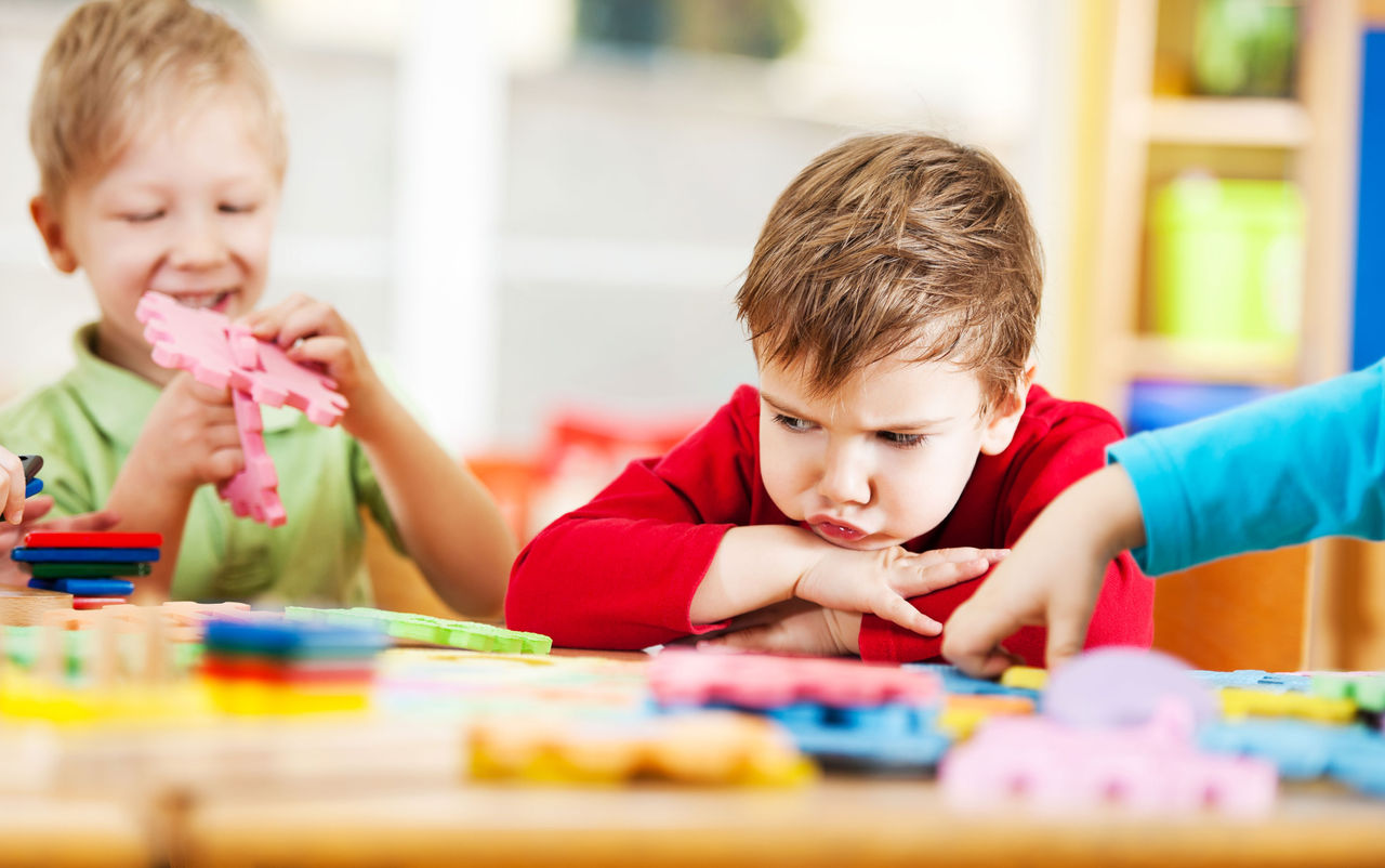 A moping child sitting at a table with toys and other children