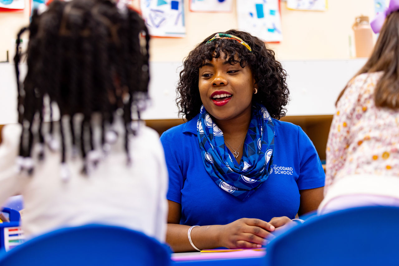 A teacher sitting at a table talking with students in a preschool classroom