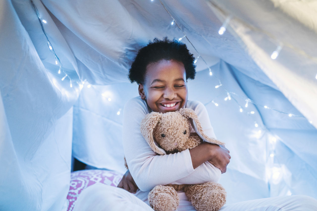 Happy girl embracing teddy bear in tent. Bed is covered with sheet. It is decorated with Christmas lights.