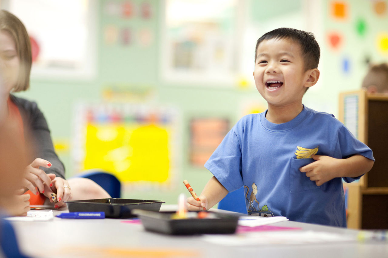 A child smiling while at a classroom table with paper and crayons