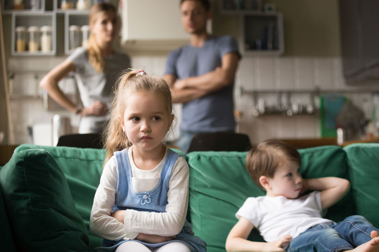two young children sitting on a couch looking upset with their parents watching on from behind