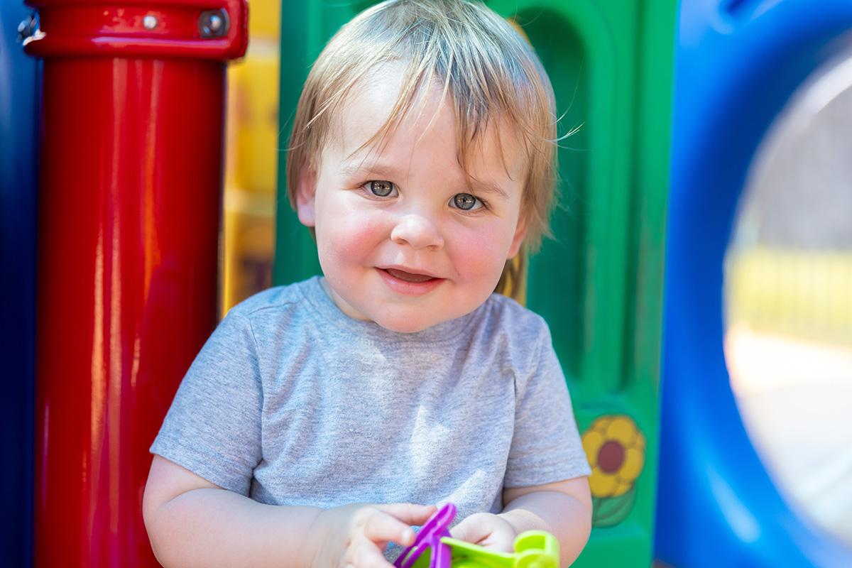 A smiling toddler with blonde hair wearing a gray shirt sits in a colorful play area. The background includes bright red, green, and blue play equipment, conveying a cheerful and playful atmosphere.
