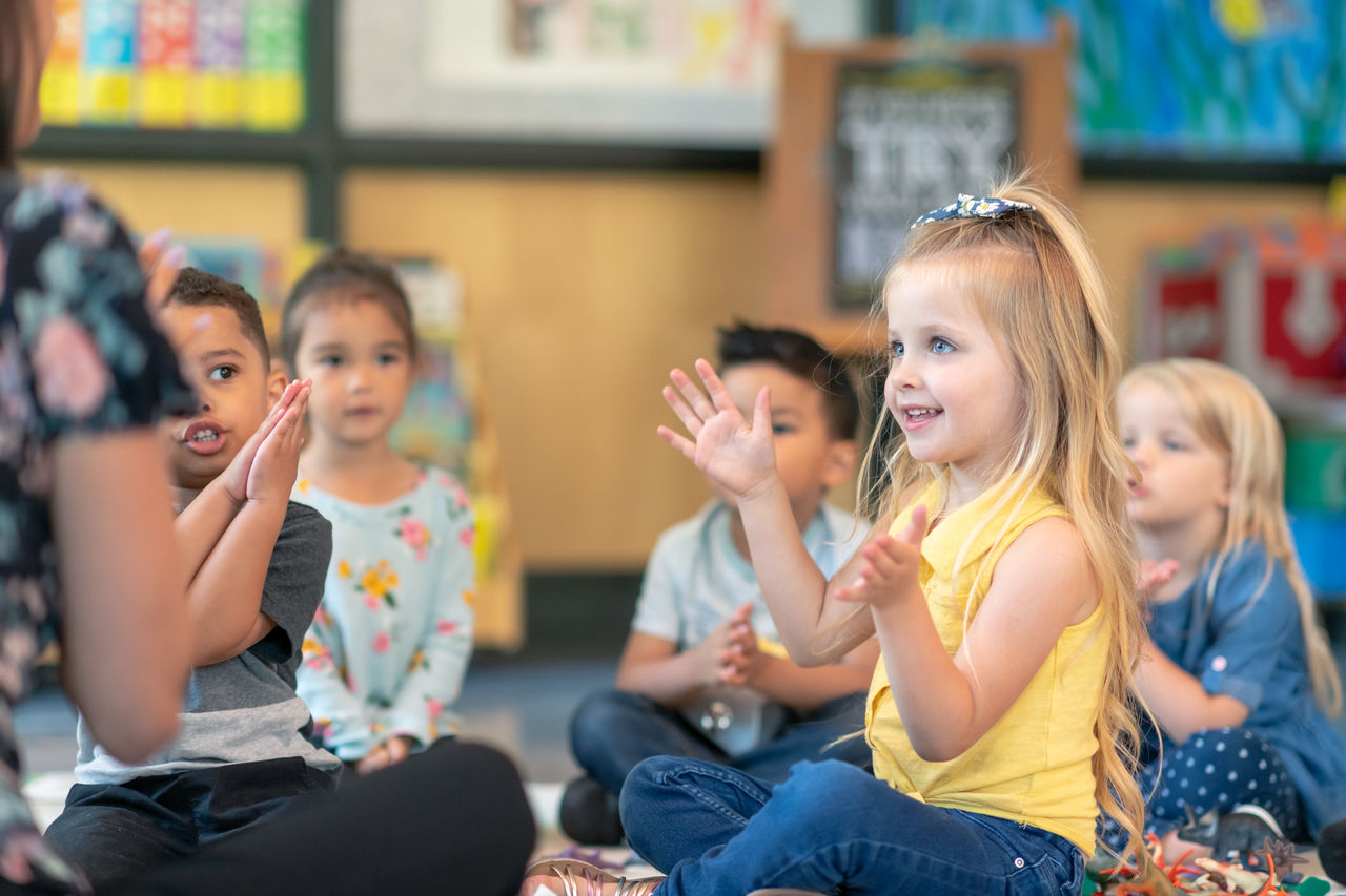 A multi-ethnic group of preschool students is sitting with their legs crossed on the floor in their classroom. The female teacher is sitting on the floor facing the children. The happy kids are smiling and following the teacher's instructions. They are clapping together. They are in music class.
