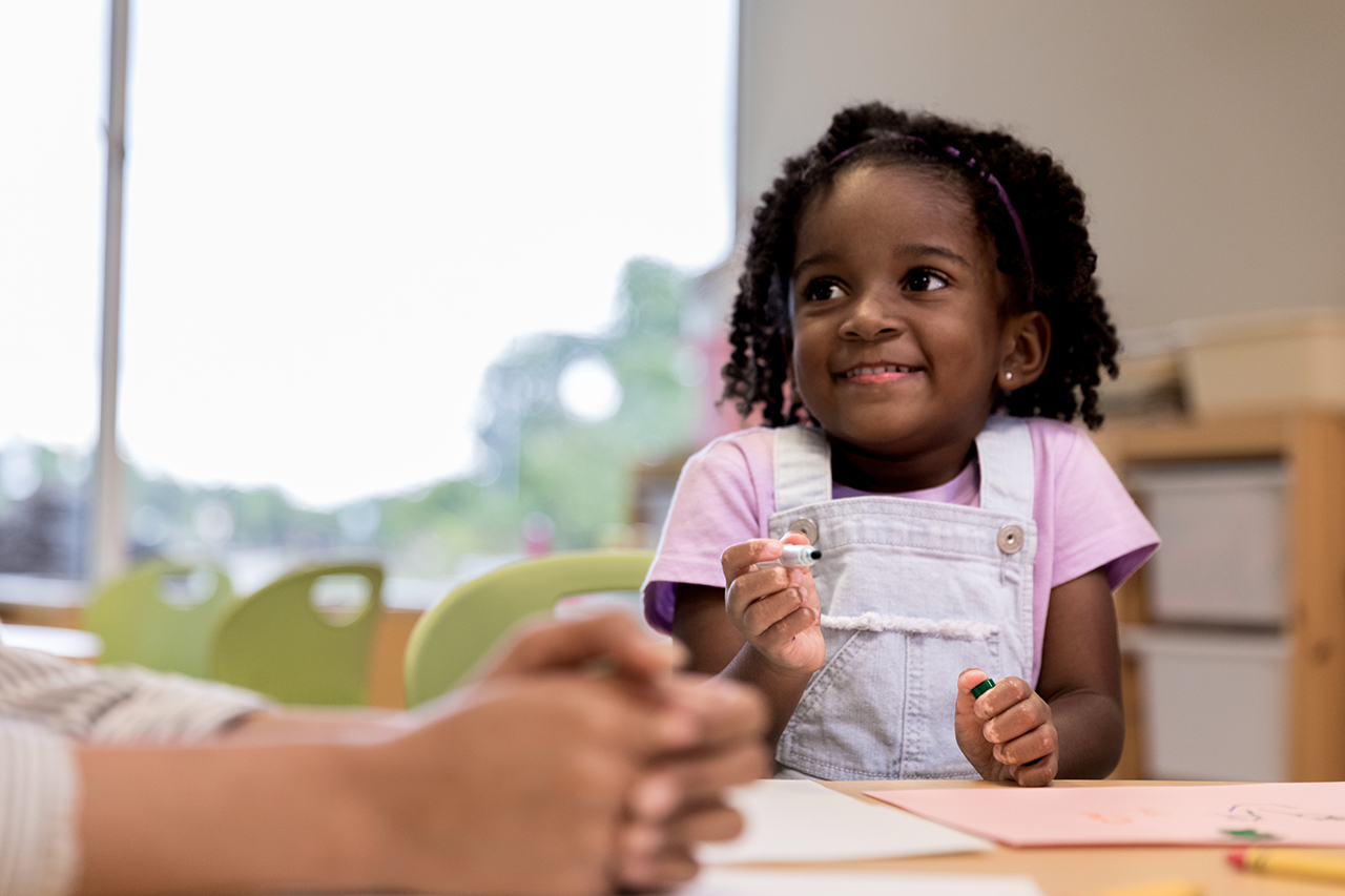 The cute young girl smiles at an unseen daycare teacher as she holds her marker.