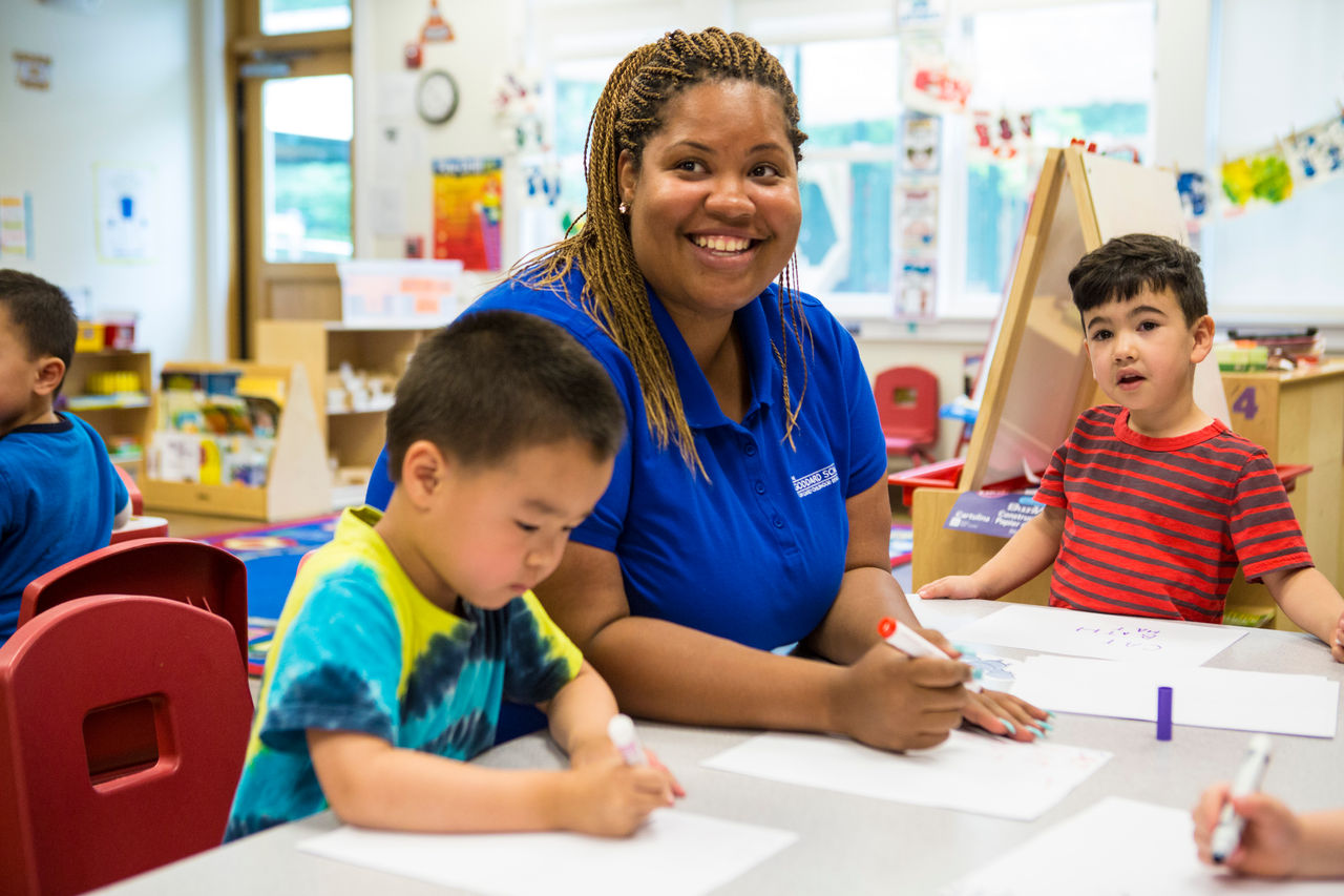 A smiling teacher draws with markers at a table alongside young children.
