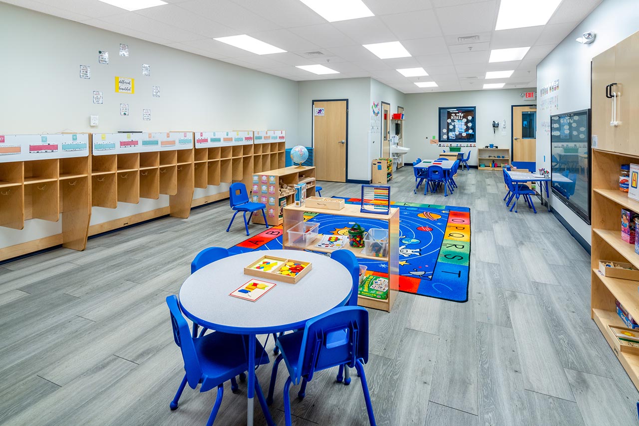 A bright, open Goddard School classroom with blue tables and chairs, hand washing station, colorful rugs and wooden bookshelves with books, toys, games and activities.