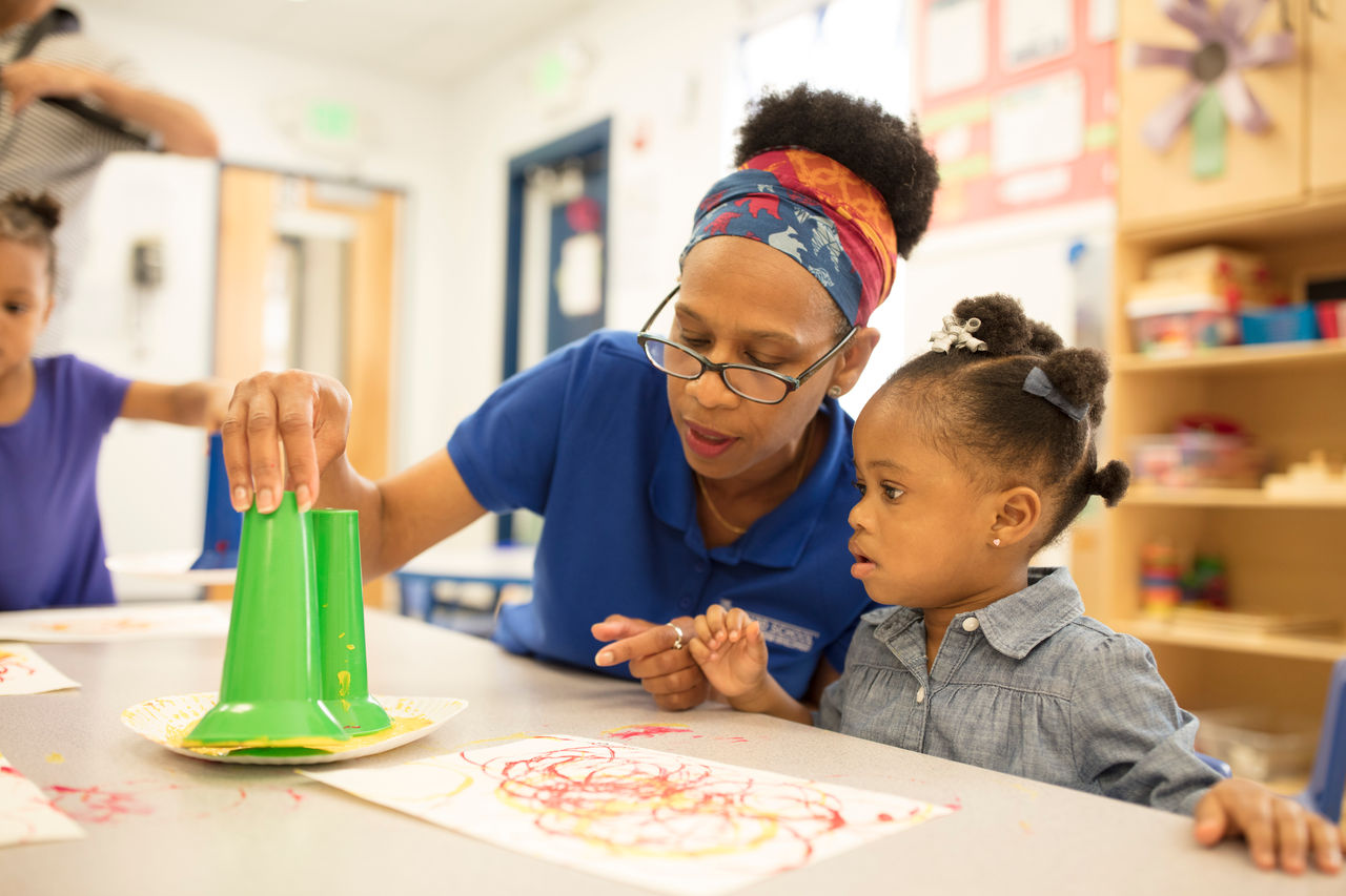 A teacher helping a child make art patterns on paper out of green funnels dipped in paint.