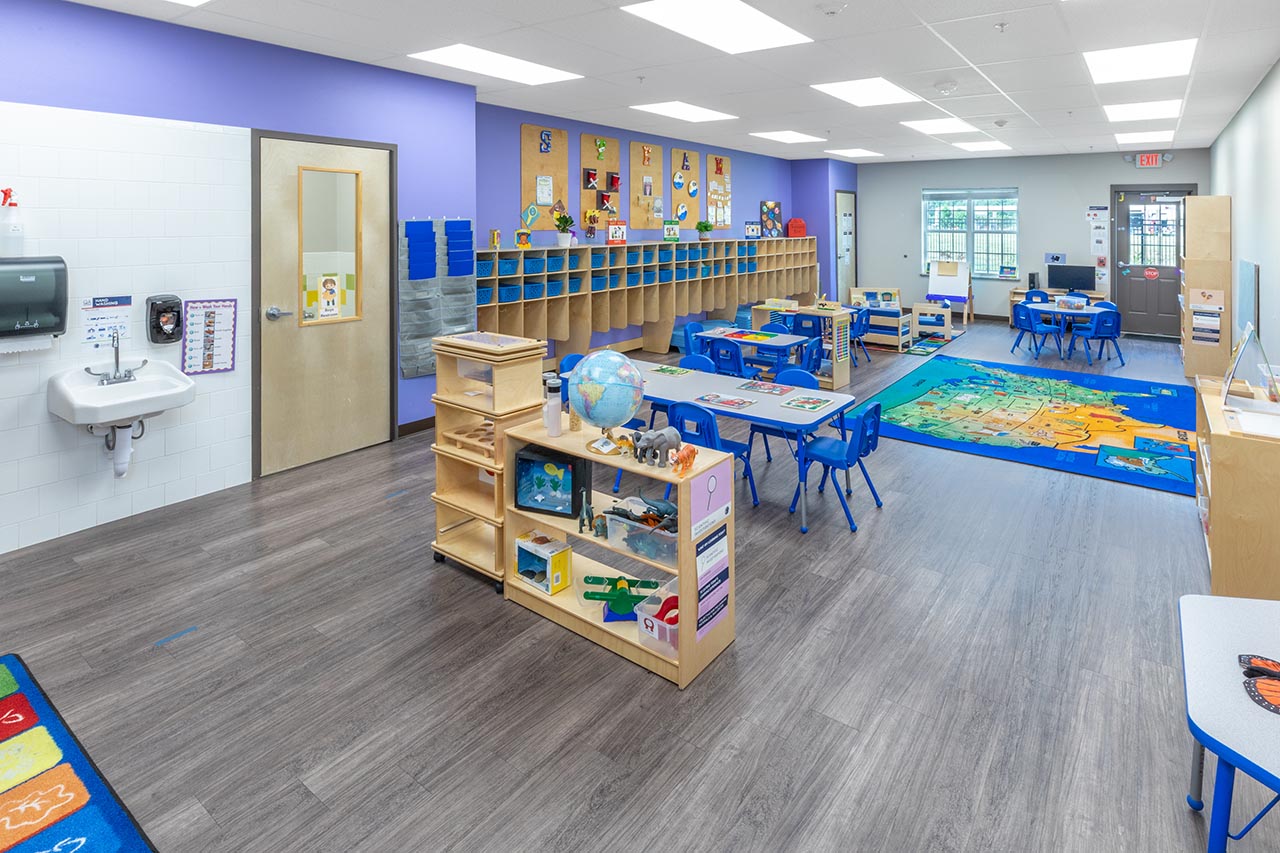 An open, bright Goddard School classroom with small green tables and chairs, wooden activity bins, a dress up corner and small cubbies. 