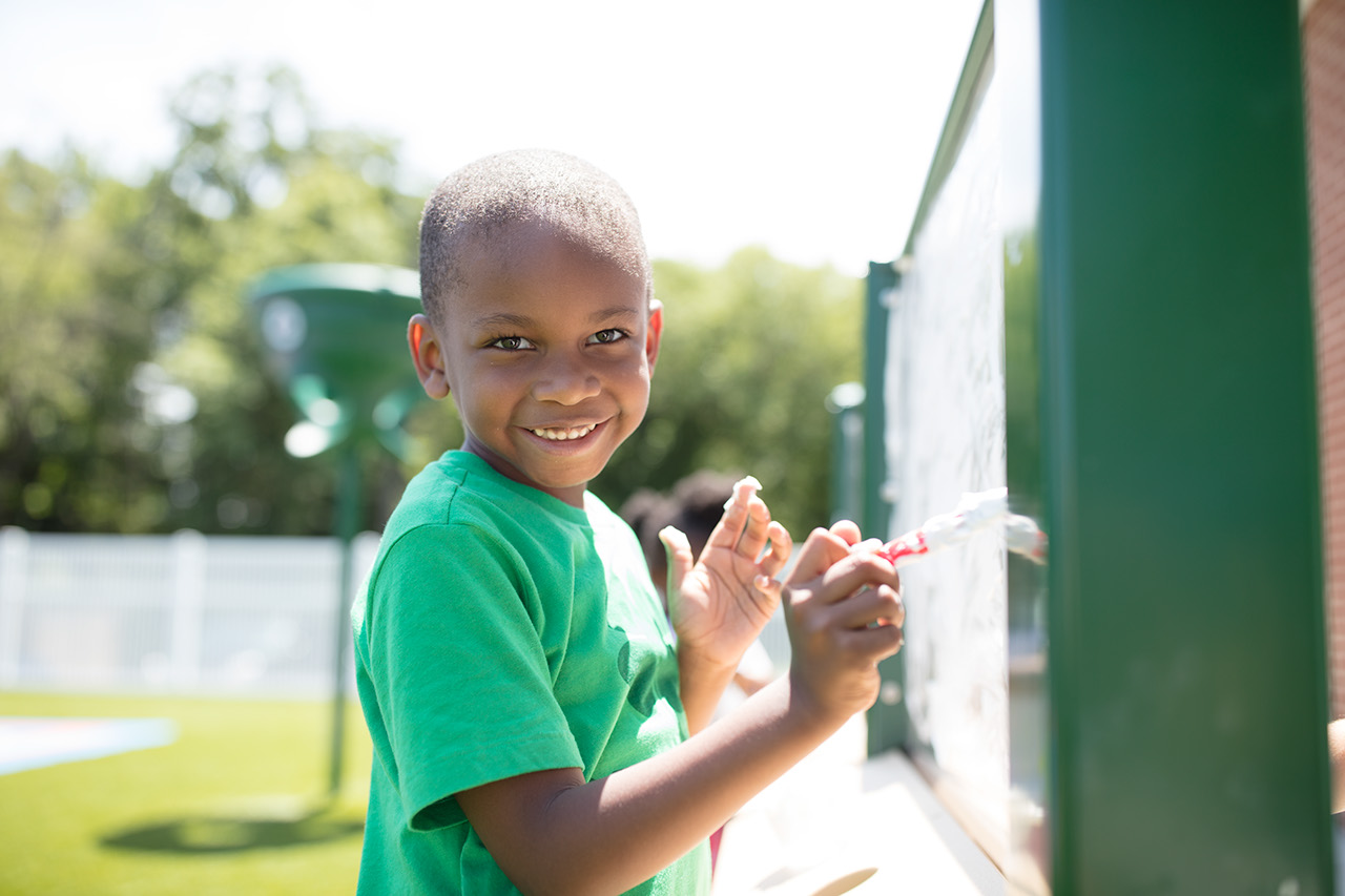 A smiling boy draws on a dry erase activity board outdoors.