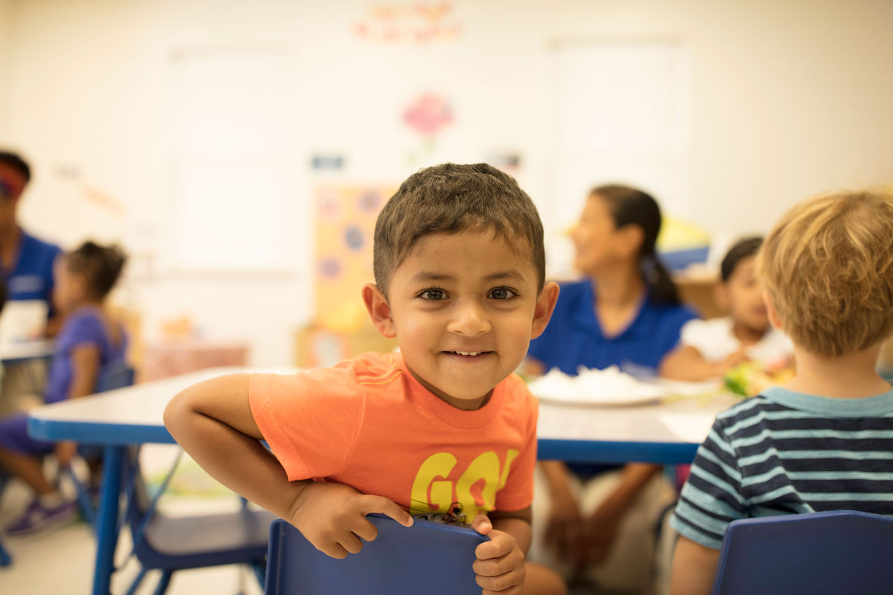 A young child inside a classroom, turned around in his chair with a big smile.