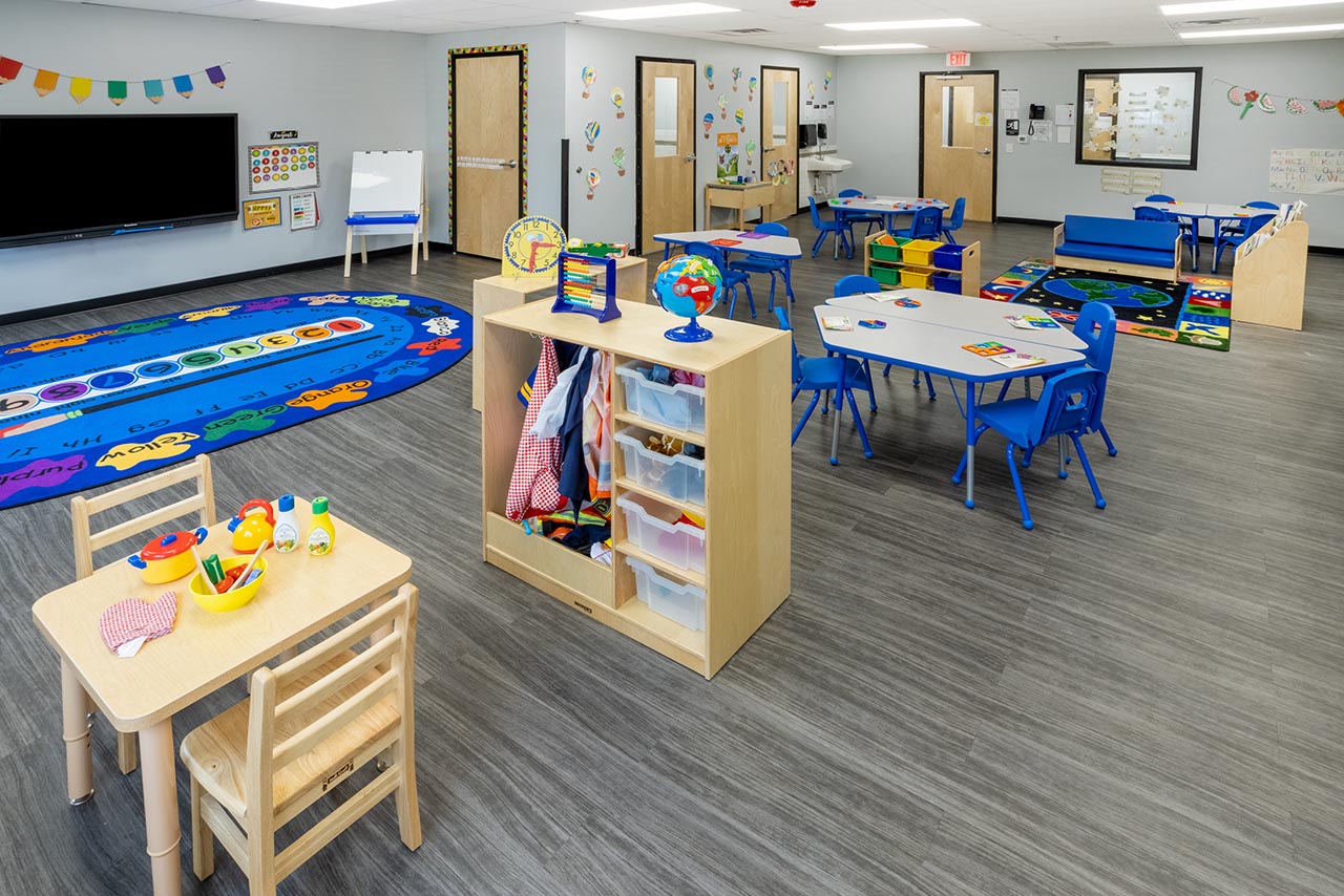 A bright, open Goddard School classroom with blue tables and chairs, hand washing station, colorful rugs and wooden bookshelves with books, toys, games and activities.