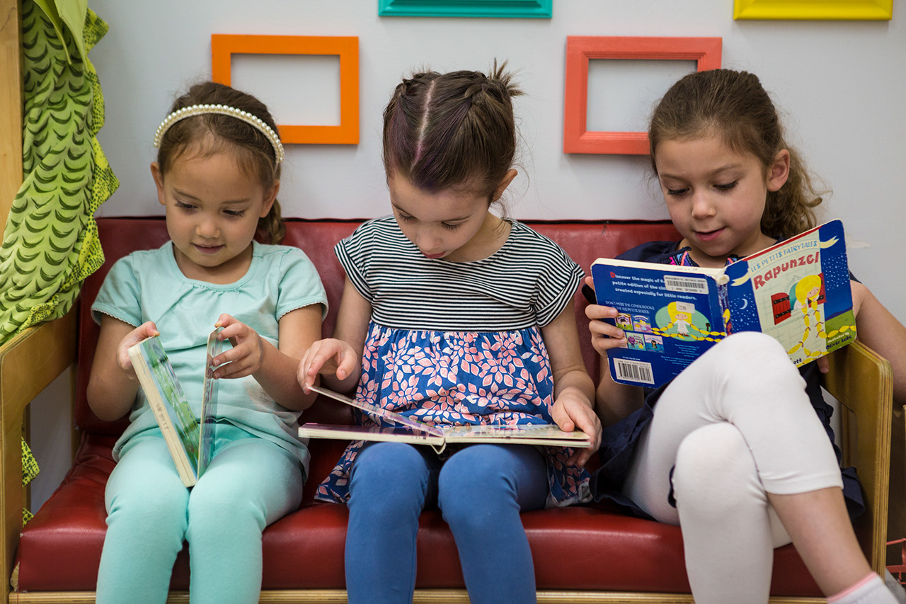 Three girls sit  on a red bench, each reading a book.
