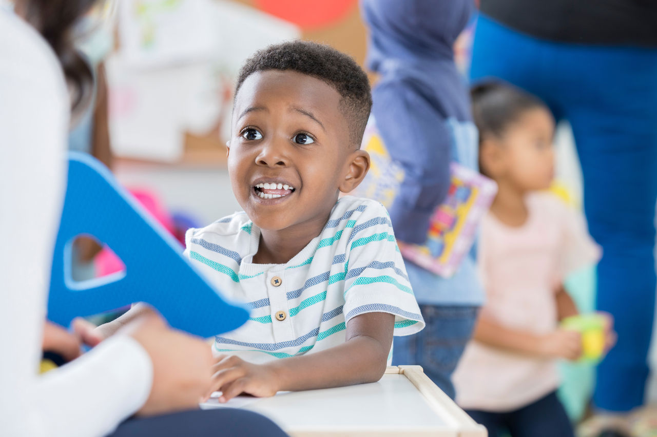An excited child sitting at a table across from a teacher who is holding up a big blue letter "A".