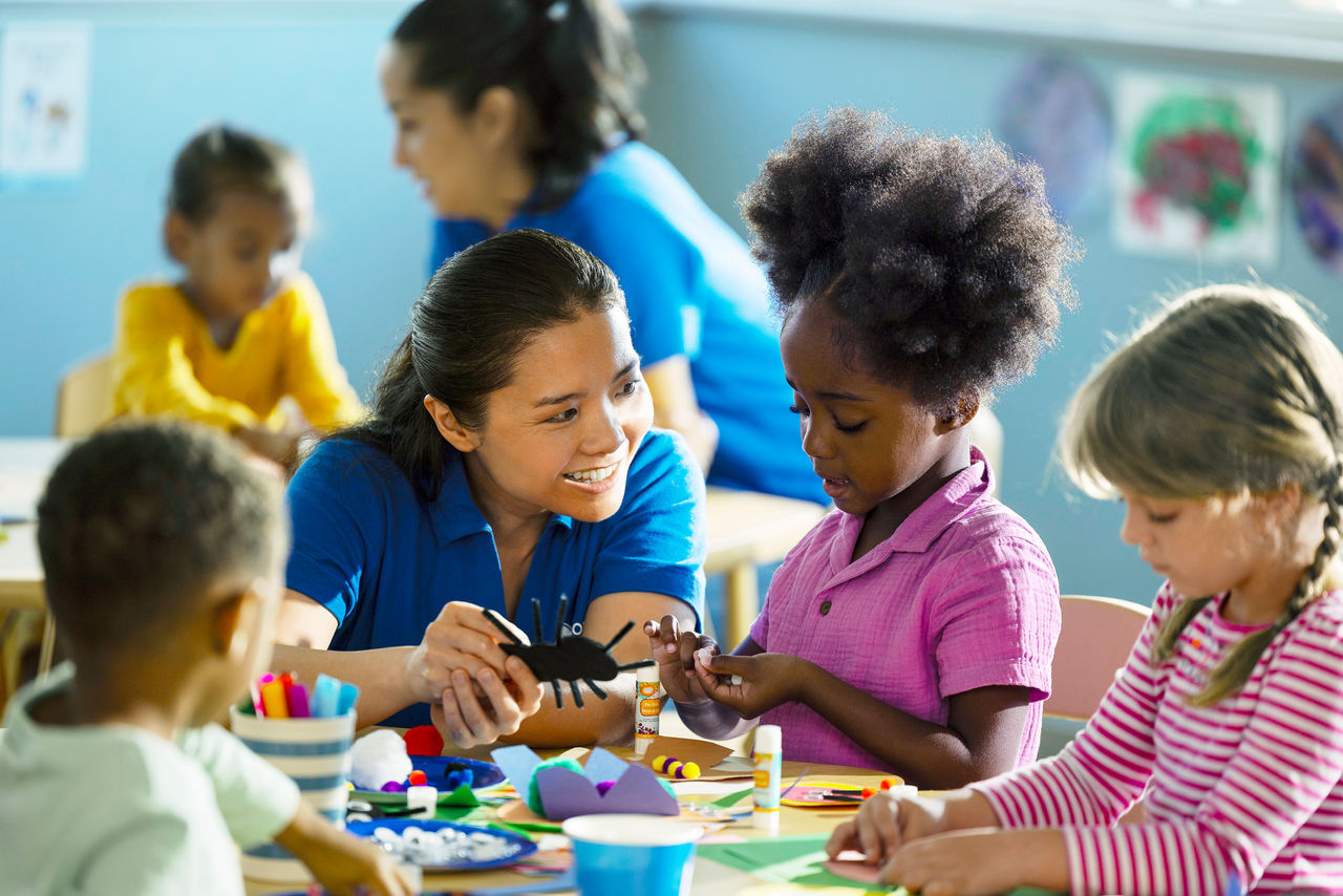 A smiling teacher holds up a lady bug to a happy child in a paint spattered shirt.