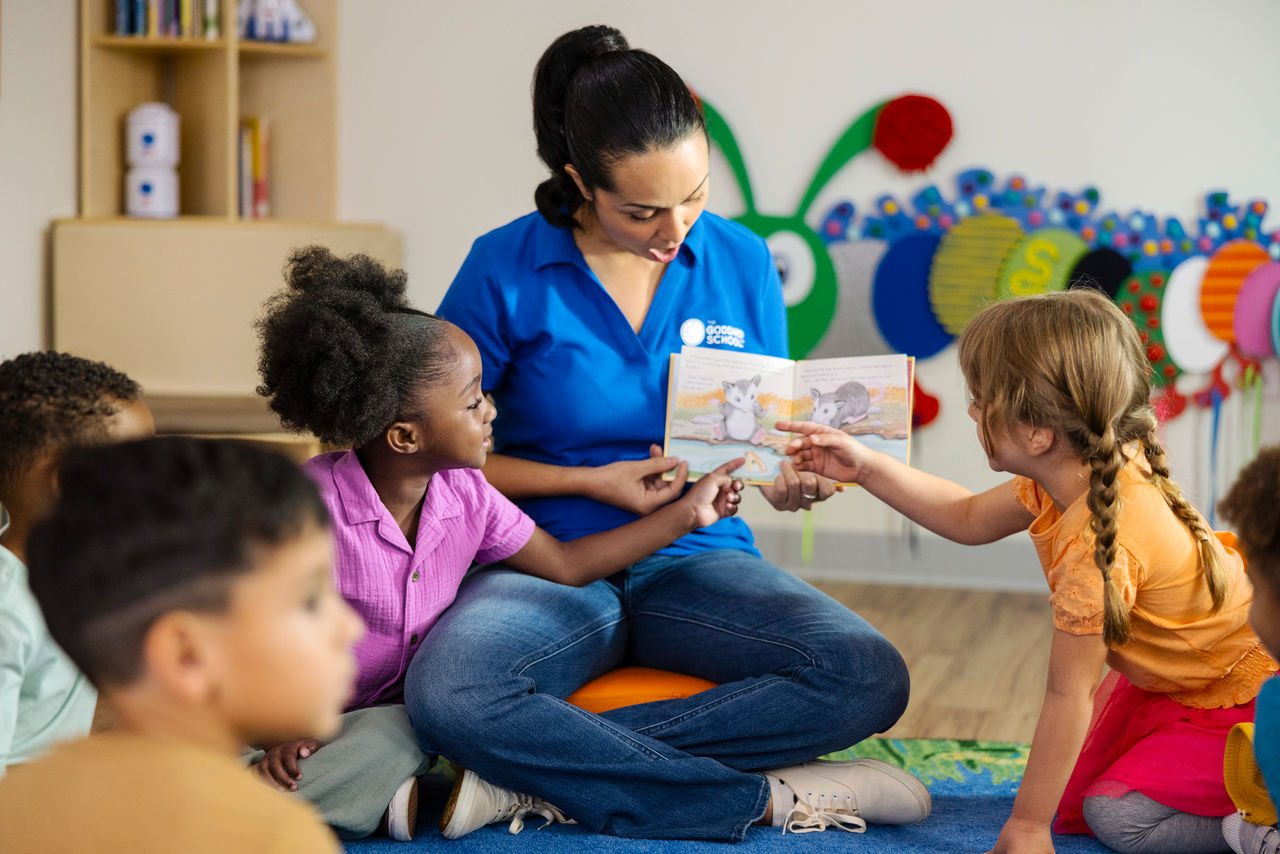 Young children point to pictures in a book a teacher is reading to children while sitting on the floor of a preschool classroom