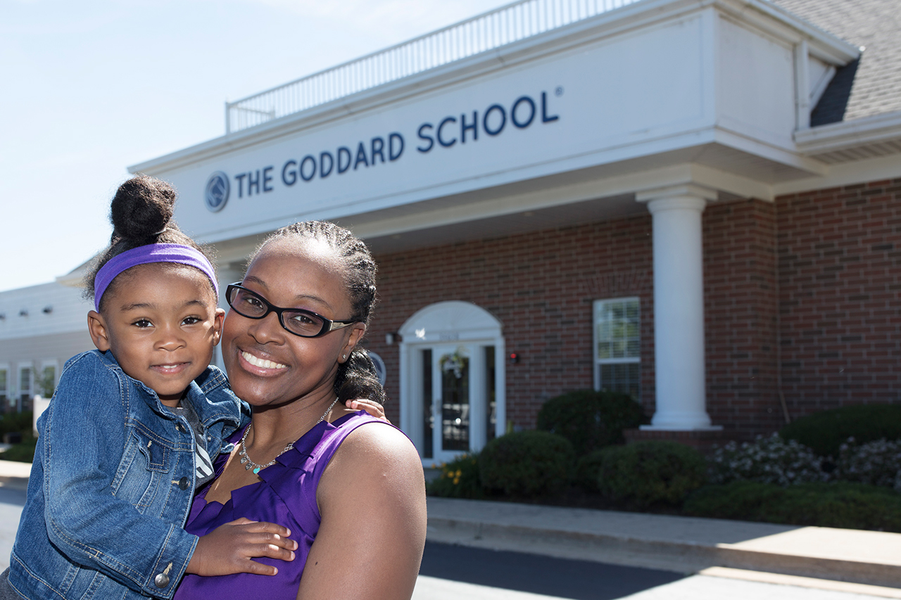 A mother and child stand outside The Goddard School on a sunny day.