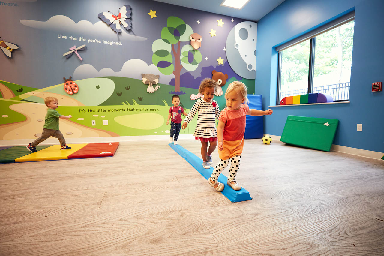 Small children walk along a low balance beam in an indoor gym.