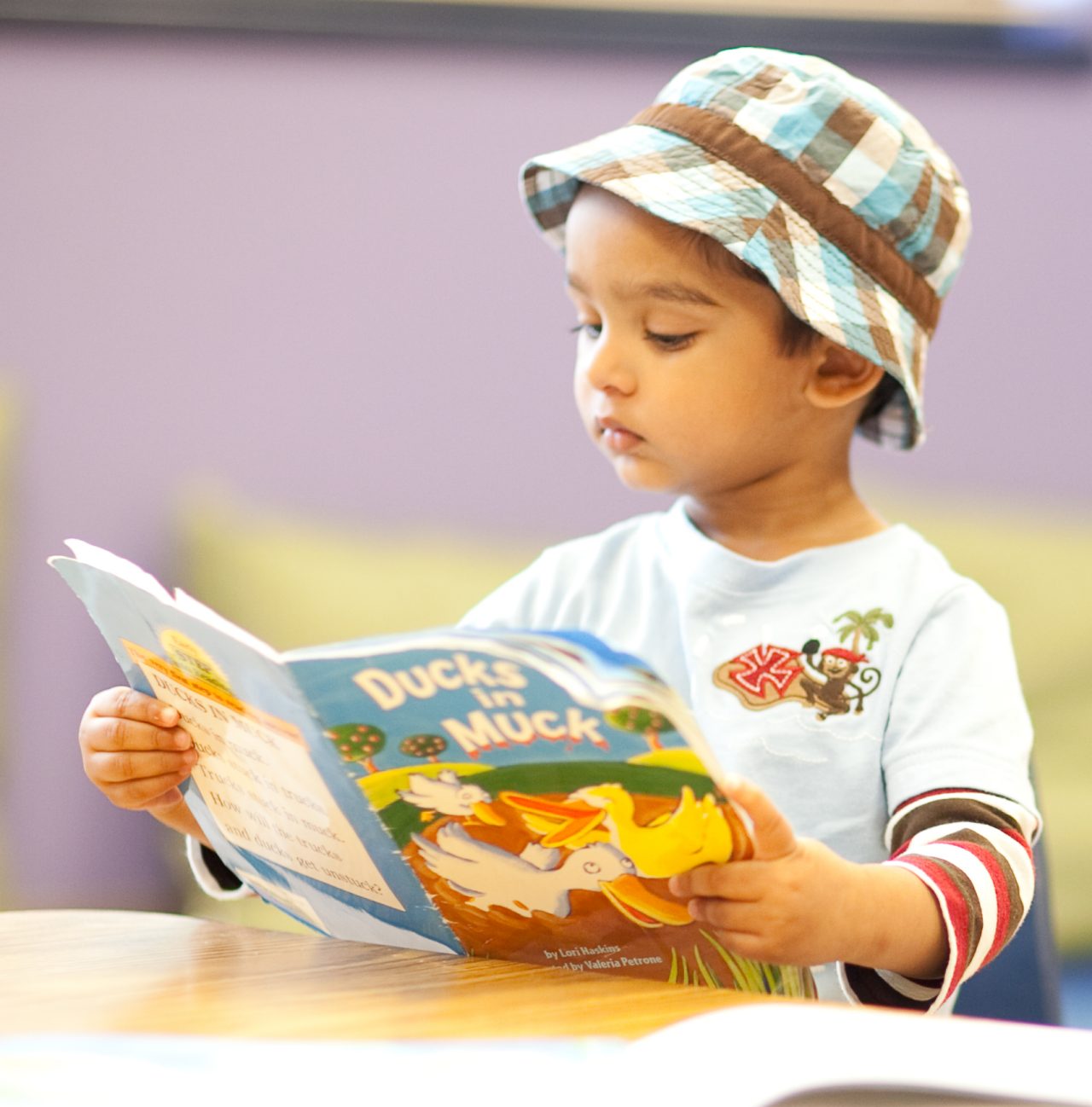 A young boy in a bucket hat reads a book, seated next to a plush monkey at a table.