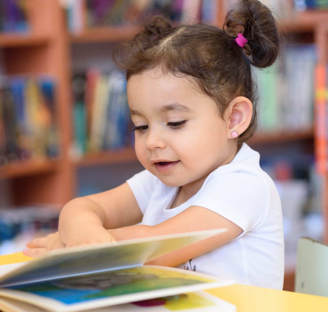 Little Girl Indoors In Front Of Books. Cute Young Toddler Sitting On A Chair Near Table and Reading Book. Child reads in a bookstore, surrounded by colorful books. Library, Shop, Shelving In Home.