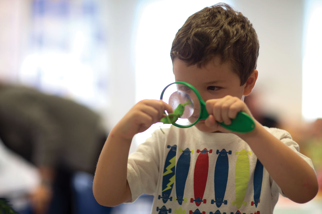 A young child looking closely at a green toy object through a green magnifying glass.