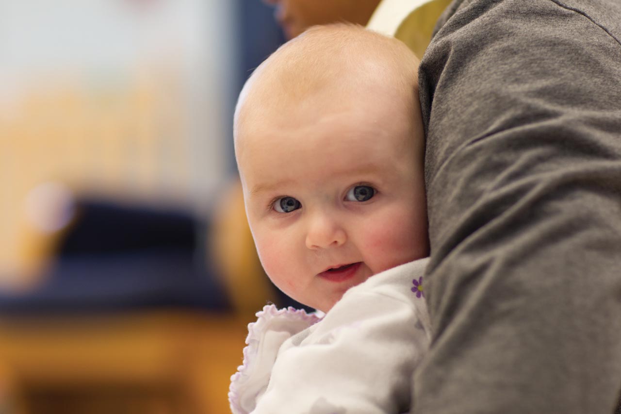 A curious baby held on a teacher's lap, looking back into the camera.