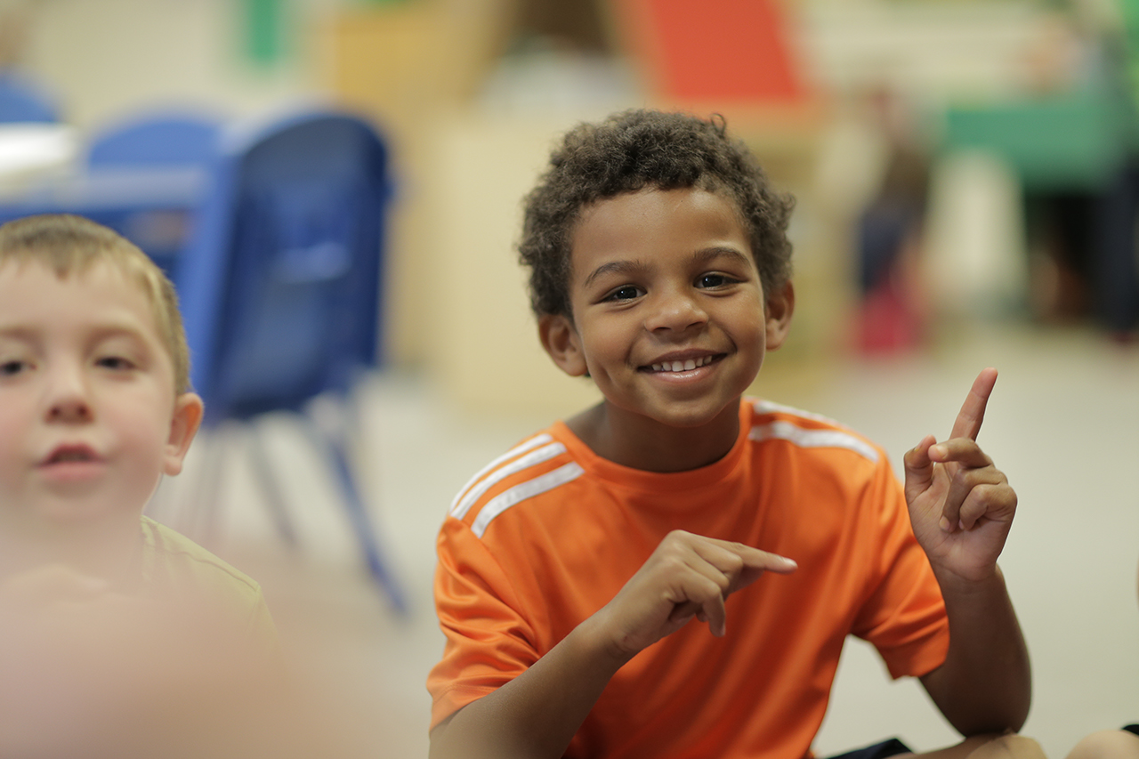 A child in an orange t-shirt sitting, smiling, and pointing both fingers.