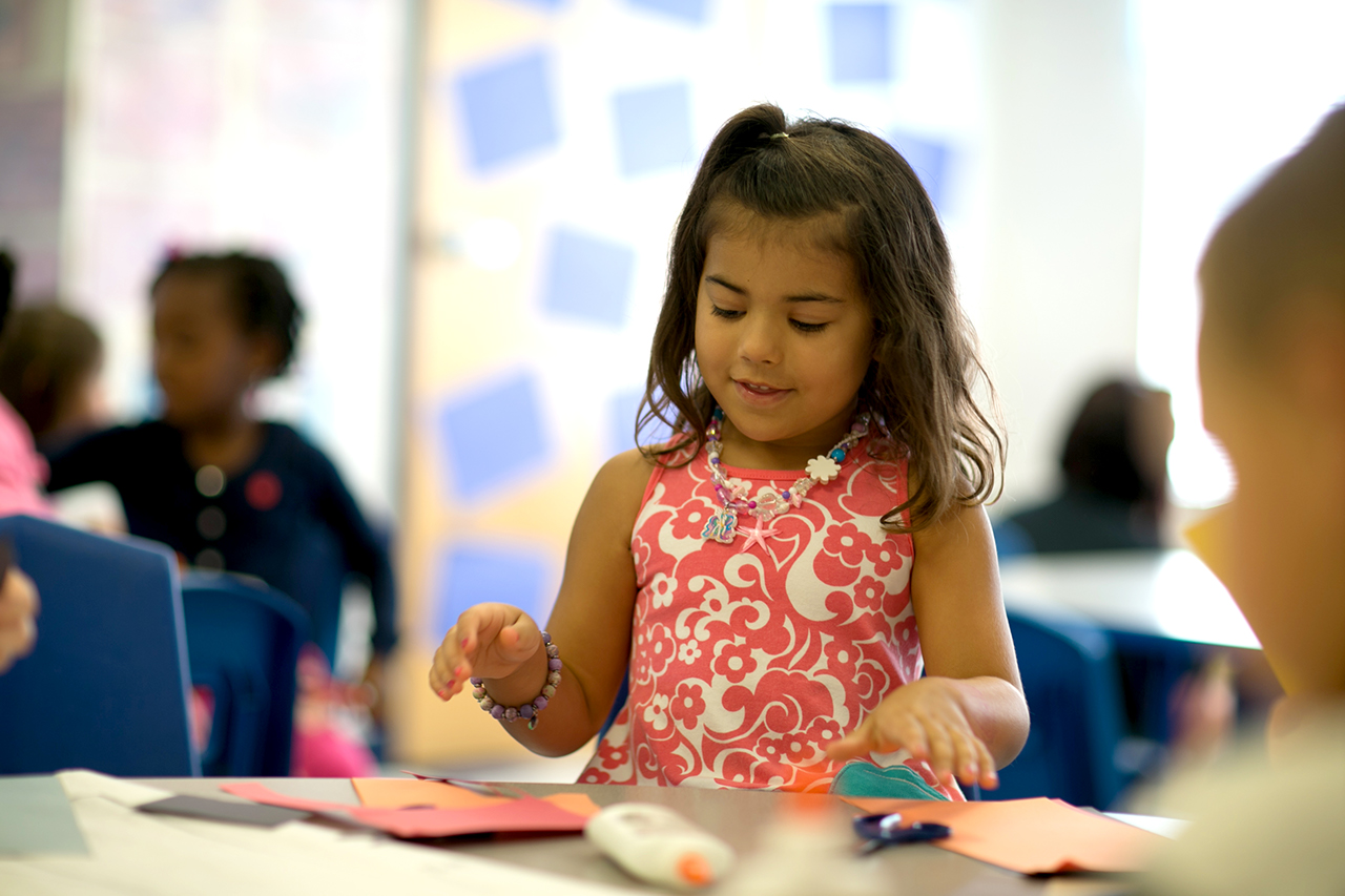 A child in a pink floral shirt sitting at a table doing arts and crafts with construction paper, glue, and safety scissors.