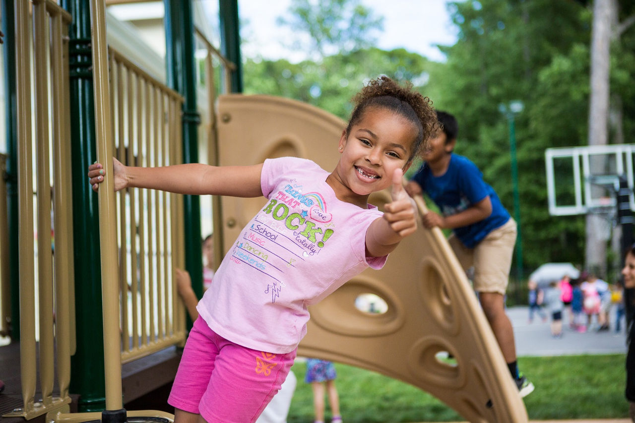 A child holding onto the bars of a play structure, leaning outward, smiling, and holding out a thumbs up.